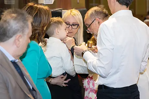Un bambino si prepara a ricevere l’unzione durante il rito del battesimo, circondato da genitori e celebrante – servizio fotografico battesimo a Milano
