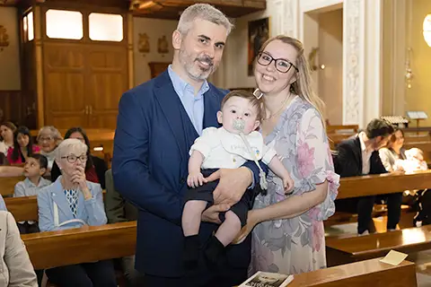 Mamma e papà sorridono tenendo in braccio il loro bambino subito dopo il battesimo, nella chiesa a Milano – servizio fotografico di famiglia