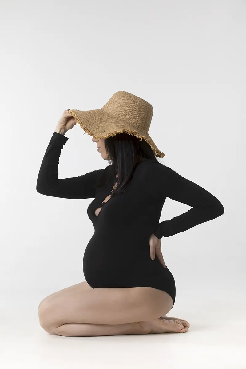 Futura mamma di profilo seduta con body nero e cappello di paglia durante un book fotografico gravidanza in studio a Milano