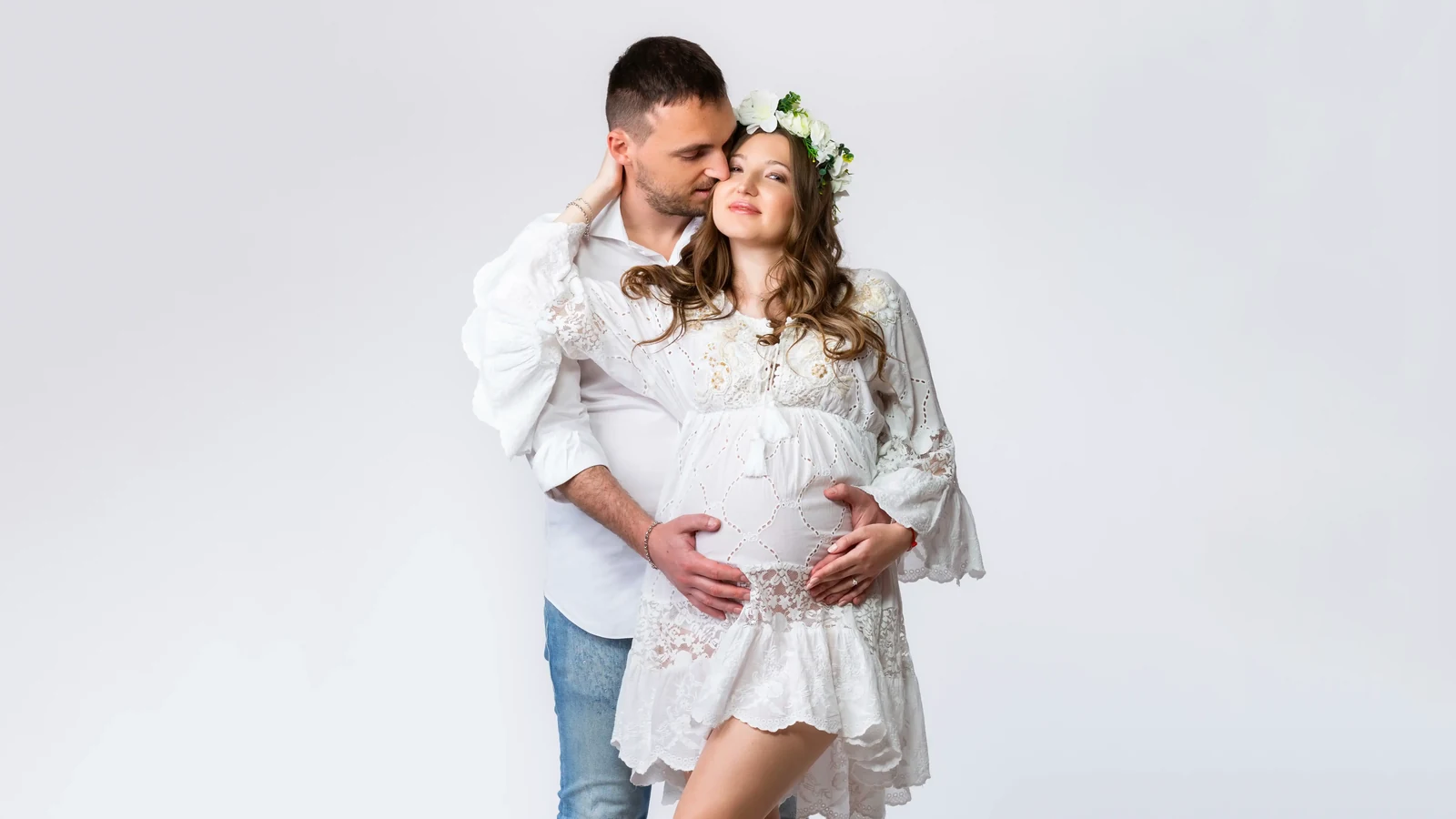 Coppia in attesa nel mio studio a Milano: lui abbraccia la futura mamma con abito bianco in pizzo e corona di fiori, mani sul pancione, book fotografico gravidanza