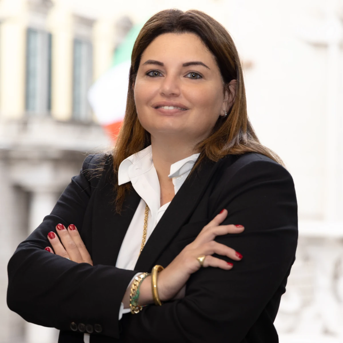 Ritratto quadrato di una donna in giacca scura e camicia bianca, braccia conserte, con sfondo urbano sfocato