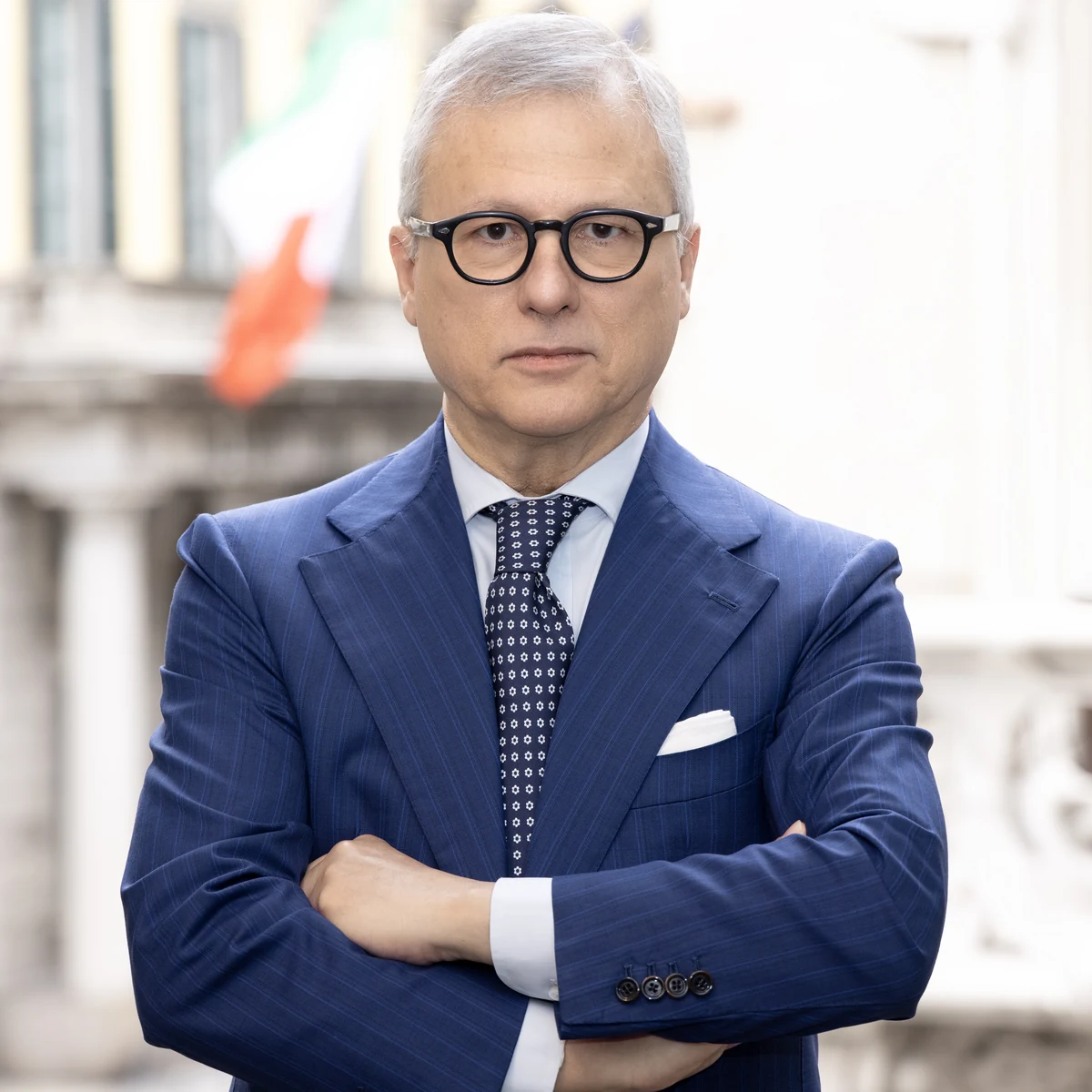 Ritratto quadrato di un uomo con occhiali e giacca blu a braccia conserte, con la città sfocata alle spalle